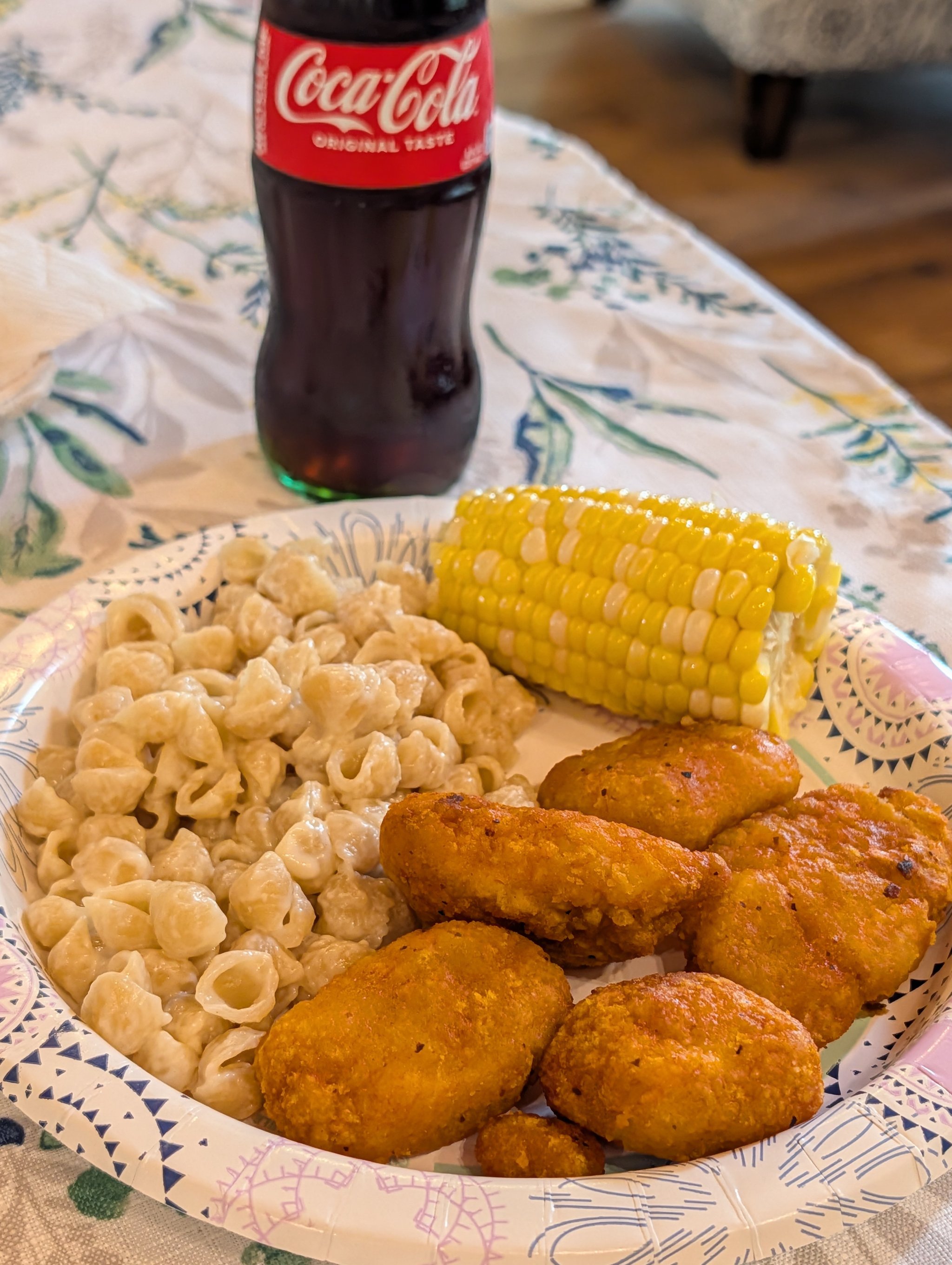 A paper plate holding a halved cob of buttered corn, creamy macaroni shells, and three pieces of fried chicken, with a glass-bottle Coca-Cola in the background on a floral tablecloth