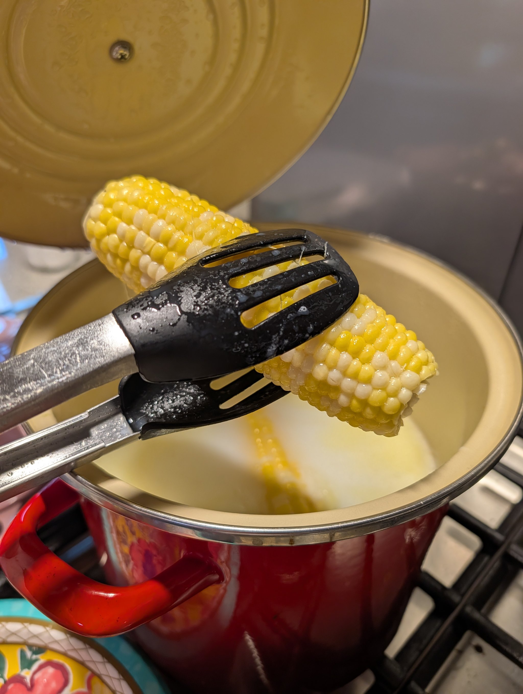 Black silicone-tipped tongs lifting two halved corn cobs out of a red Dutch oven on a gas stovetop
