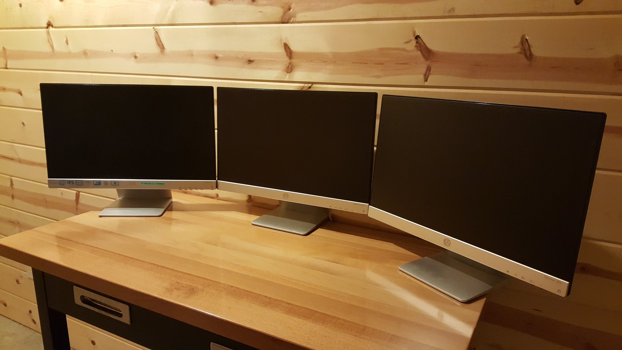 Triple monitor desk setup on a wood desk in a cabin, showing three matching displays side by side