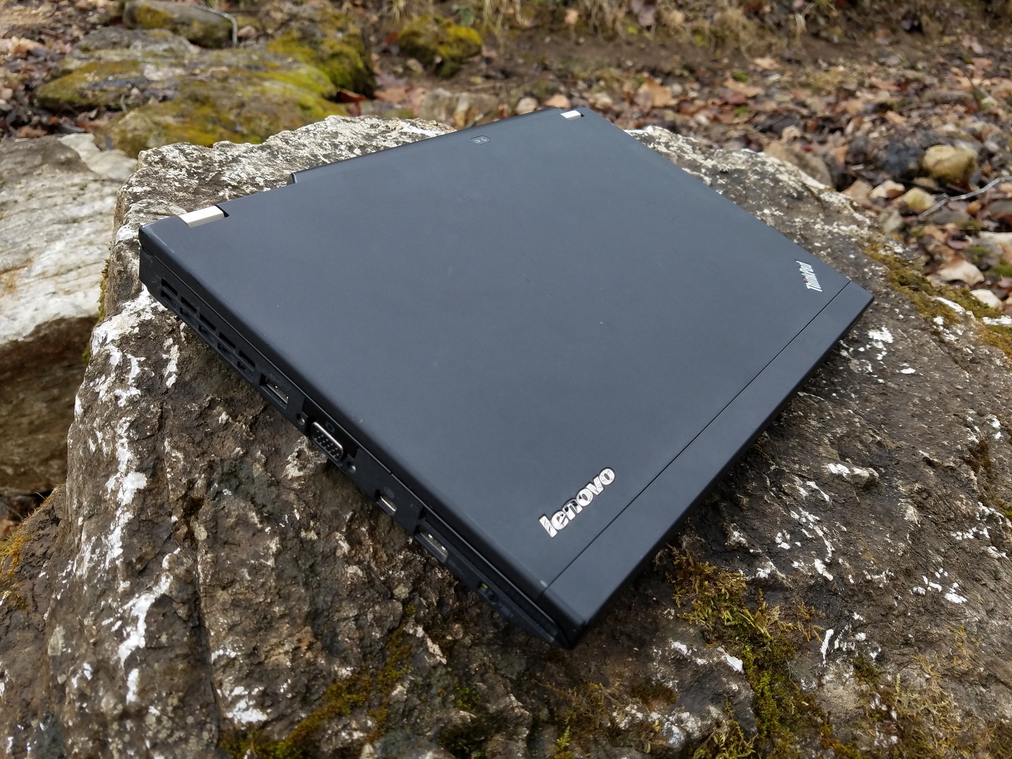 Closed Lenovo-era ThinkPad on a mossy rock outdoors. The tilted ThinkPad corner mark sits in the upper-right of the lid and a separate Lenovo wordmark sits on the lower-right of the lid. No IBM badge is present.
