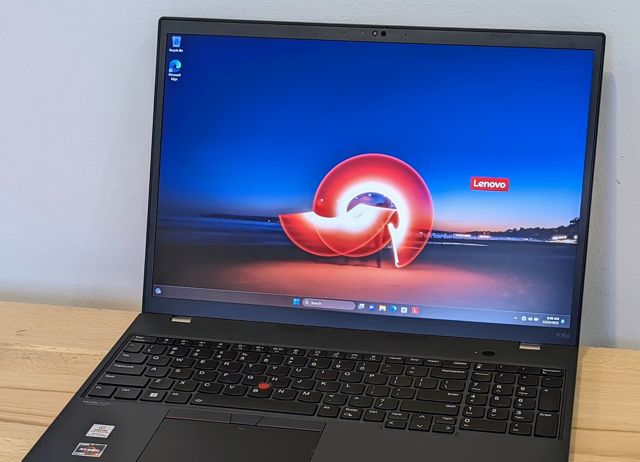 Lenovo ThinkPad P16s Gen 1 open on a wood desk. The lid is raised to display the Windows desktop with a Lenovo wallpaper. The P16s bezel label is visible at the lower-right of the screen. The keyboard, red TrackPoint, and AMD Ryzen / AMD PRO palm-rest stickers are in frame.
