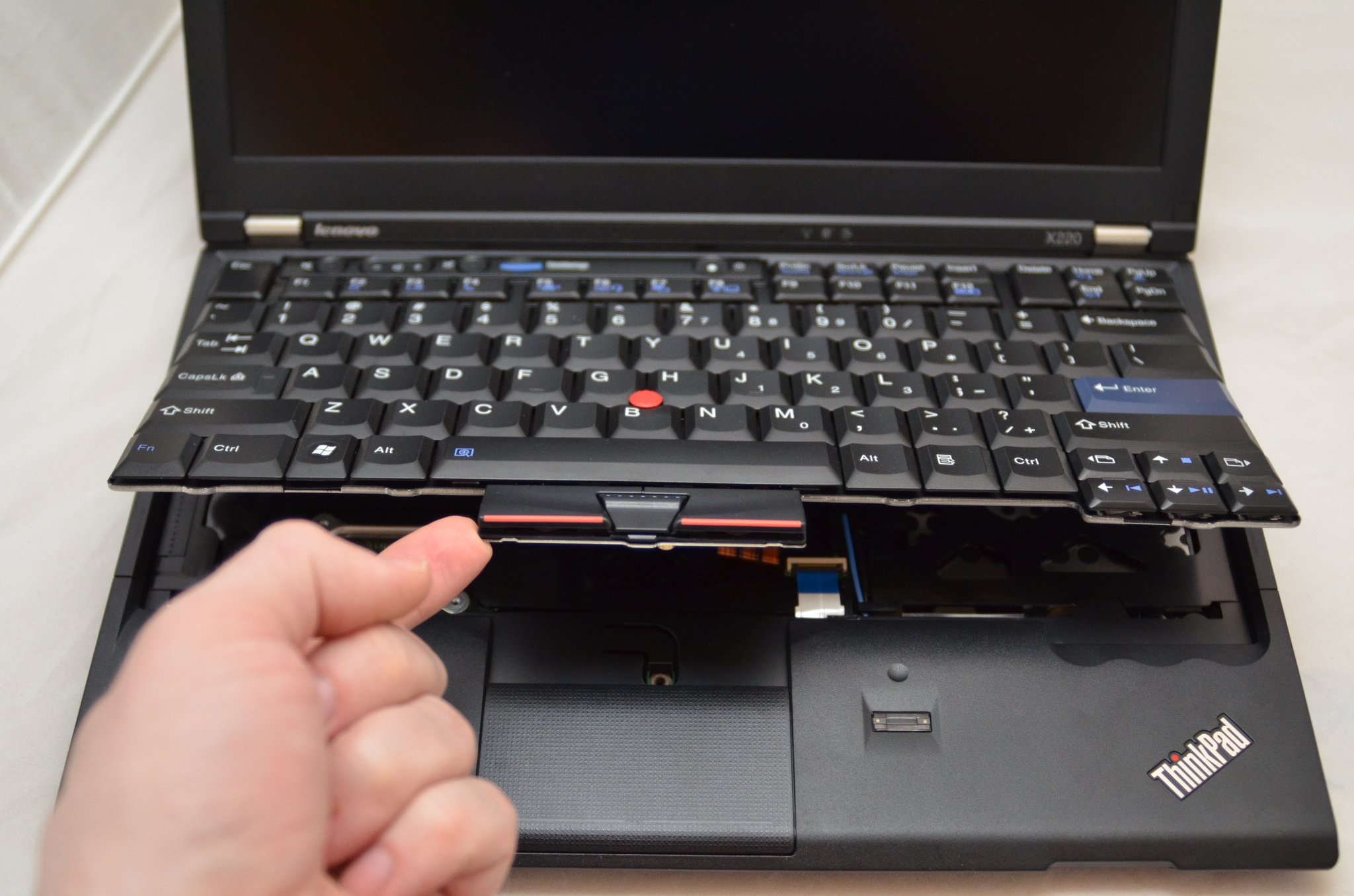ThinkPad X220 with the 7-row classic keyboard lifted out by hand mid-disassembly, showing the keyboard's plastic frame and the underlying chassis. Red TrackPoint and PageUp/PageDown cluster visible on the keyboard.
