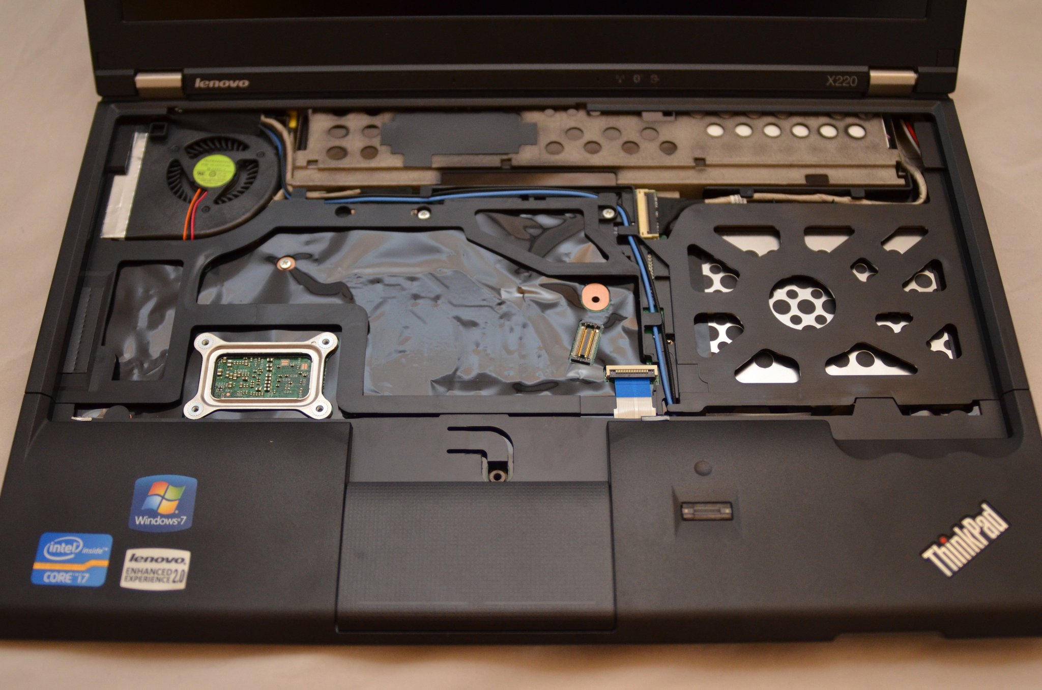 X220 chassis with the 7-row keyboard removed. Visible: the CPU cooling fan and copper heat pipe in the upper-left corner, the TrackPoint daughter-board on the left under where the spacebar sat, the silver heat-shielding plate covering most of the motherboard center, a blue dock-connector ribbon cable on the right, and Windows 7 / Intel Core i7 / Lenovo Enhanced Experience stickers on the palm rest.